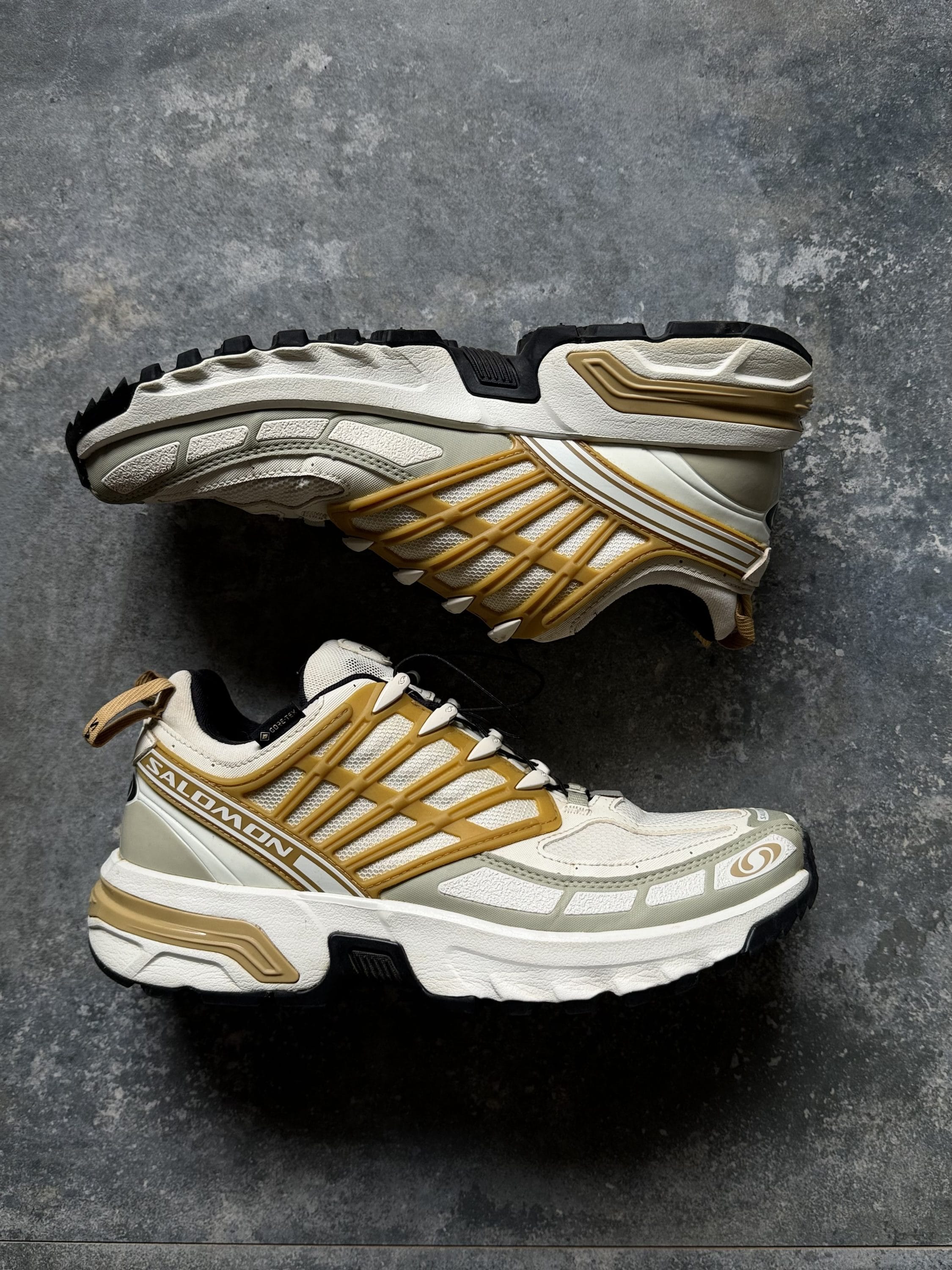 Pair of white and beige Salomon trail running shoes with gold accents photographed from side on a textured gray surface, showing rugged outsole.