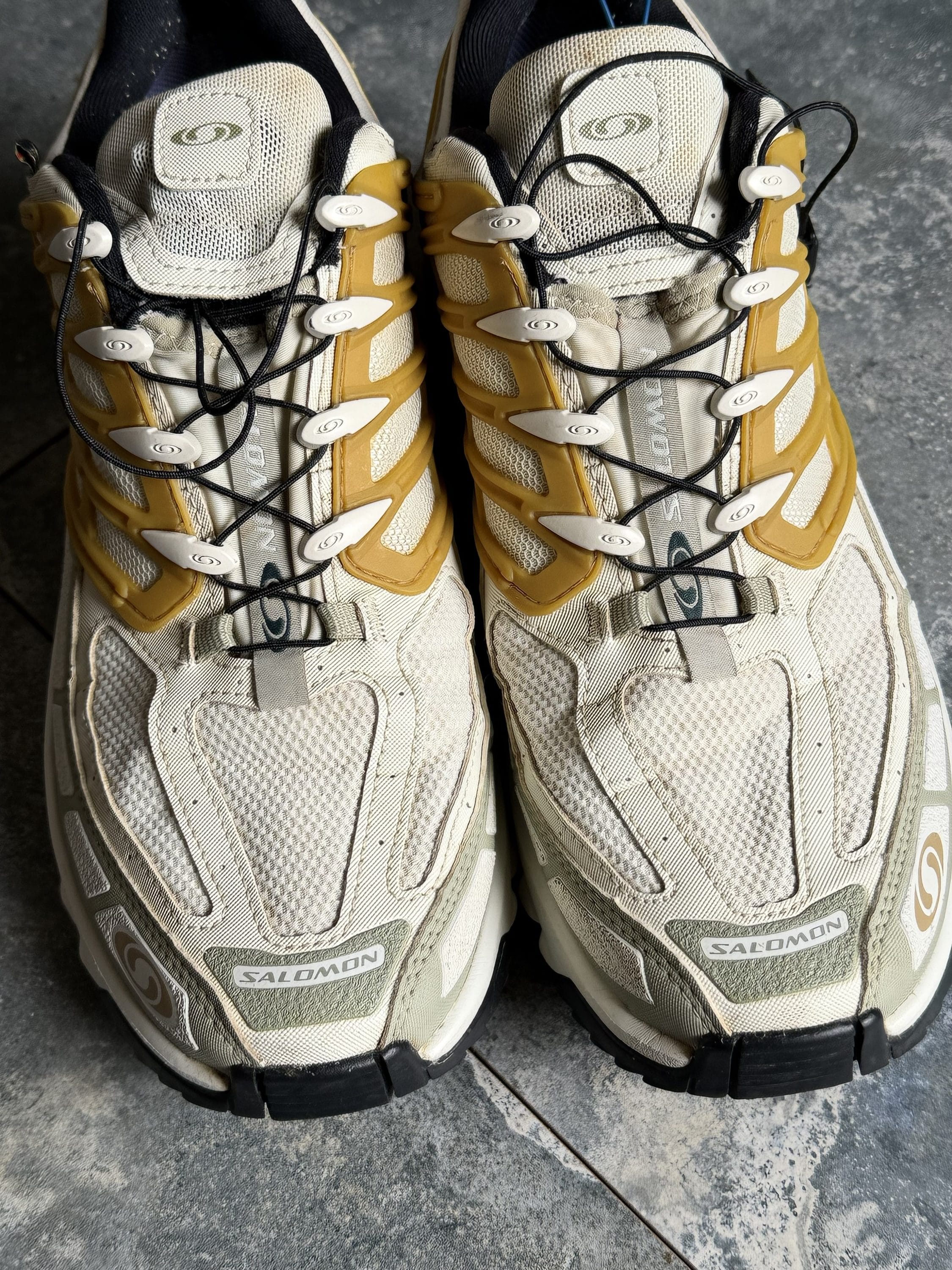 Pair of white and tan Salomon trail running shoes placed side by side on a gray concrete floor with black laces.