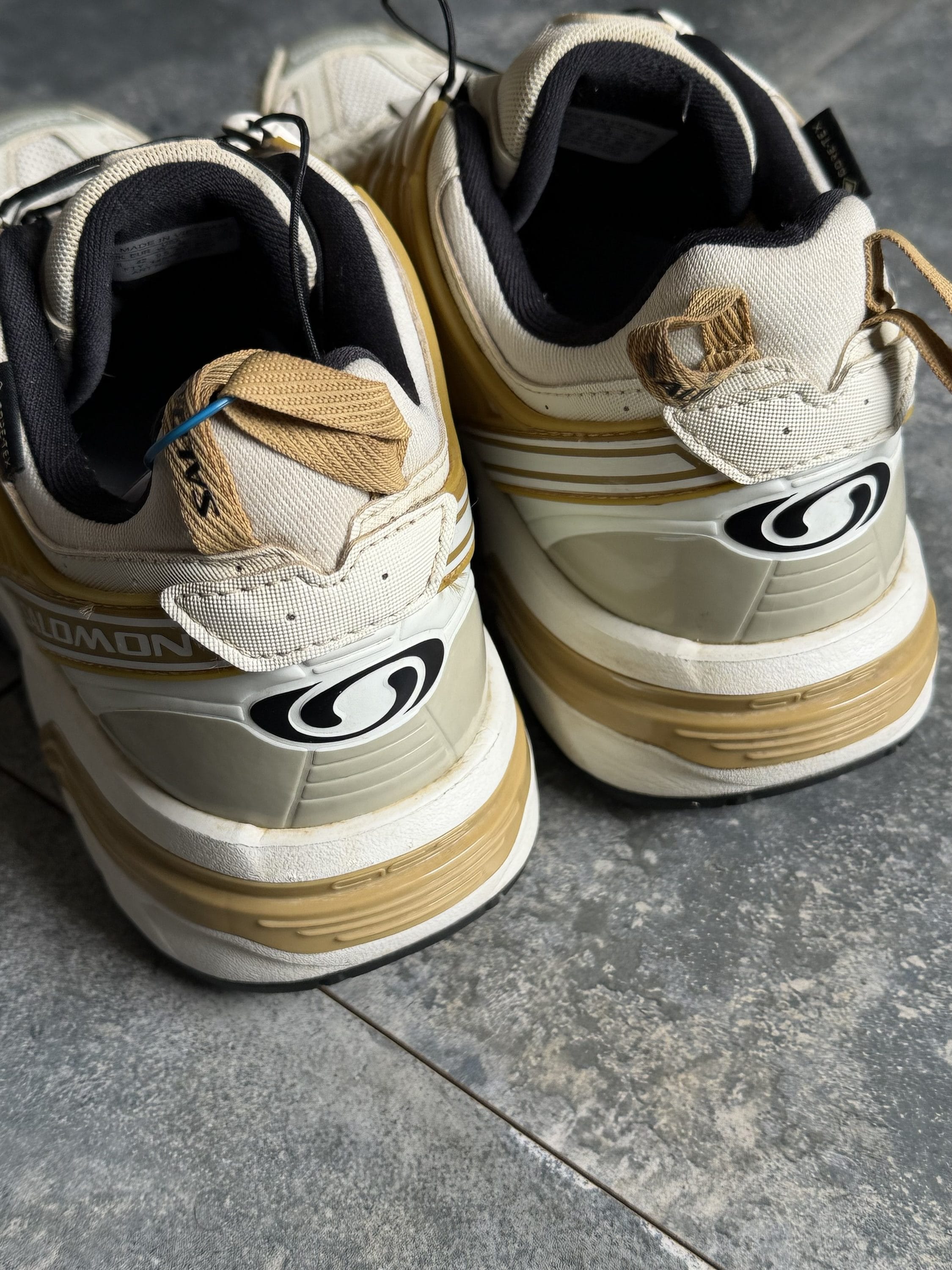 Beige sneakers with tan laces and gum soles, viewed from the back showing the Salomon logo on the heel.
