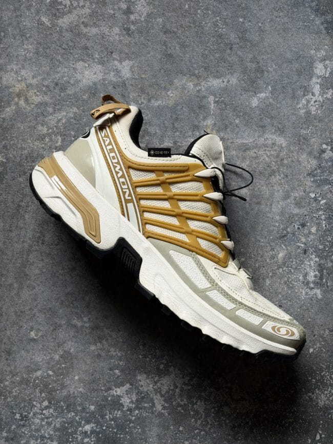 Salomon trail running shoe shown at an angle on a textured gray concrete floor, beige and white with supportive overlays