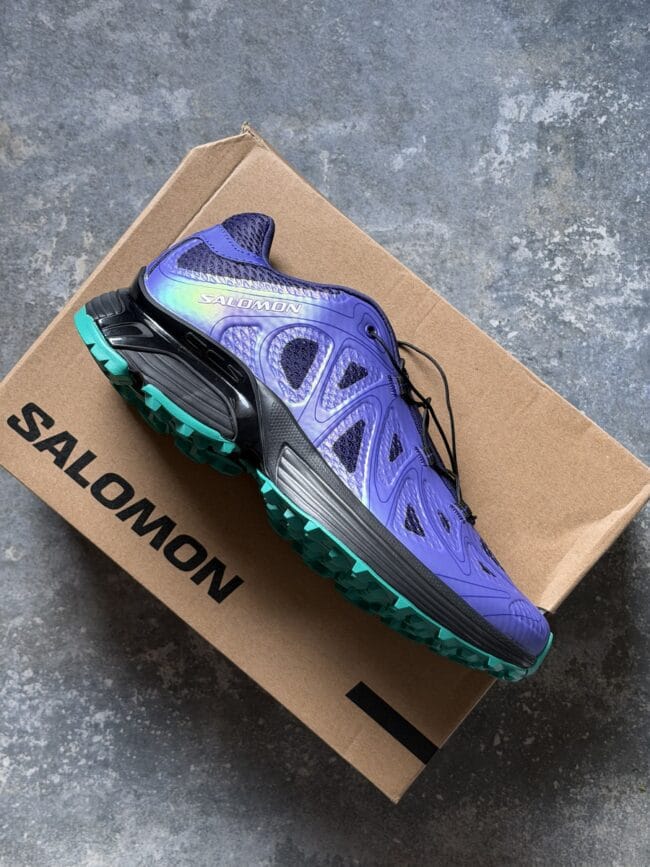 Purple Salomon trail running shoe with teal outsole resting on a brown cardboard box against a gray textured floor.