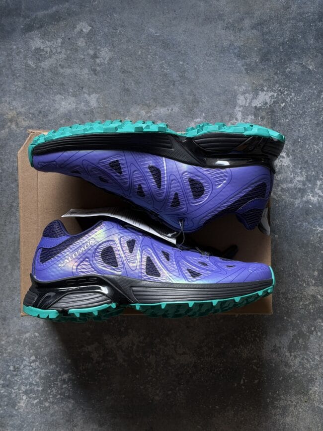 Purple Salomon trail running shoe with teal sole on a cardboard box, top-down view on a concrete floor.