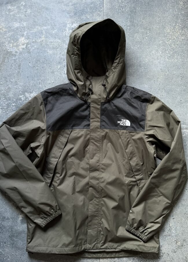 Olive-green The North Face hooded waterproof jacket laid flat on a textured surface with a black chest panel and drawstrings on the hood.