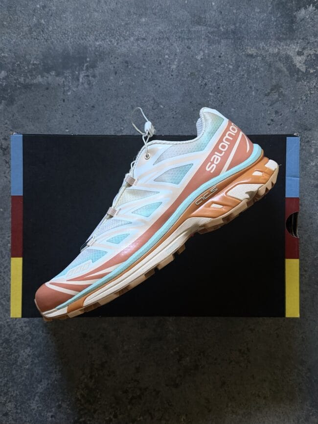 Pastel Salomon running shoe in blue, peach and cream rests diagonally on a black shoebox with blue edges on a concrete floor.