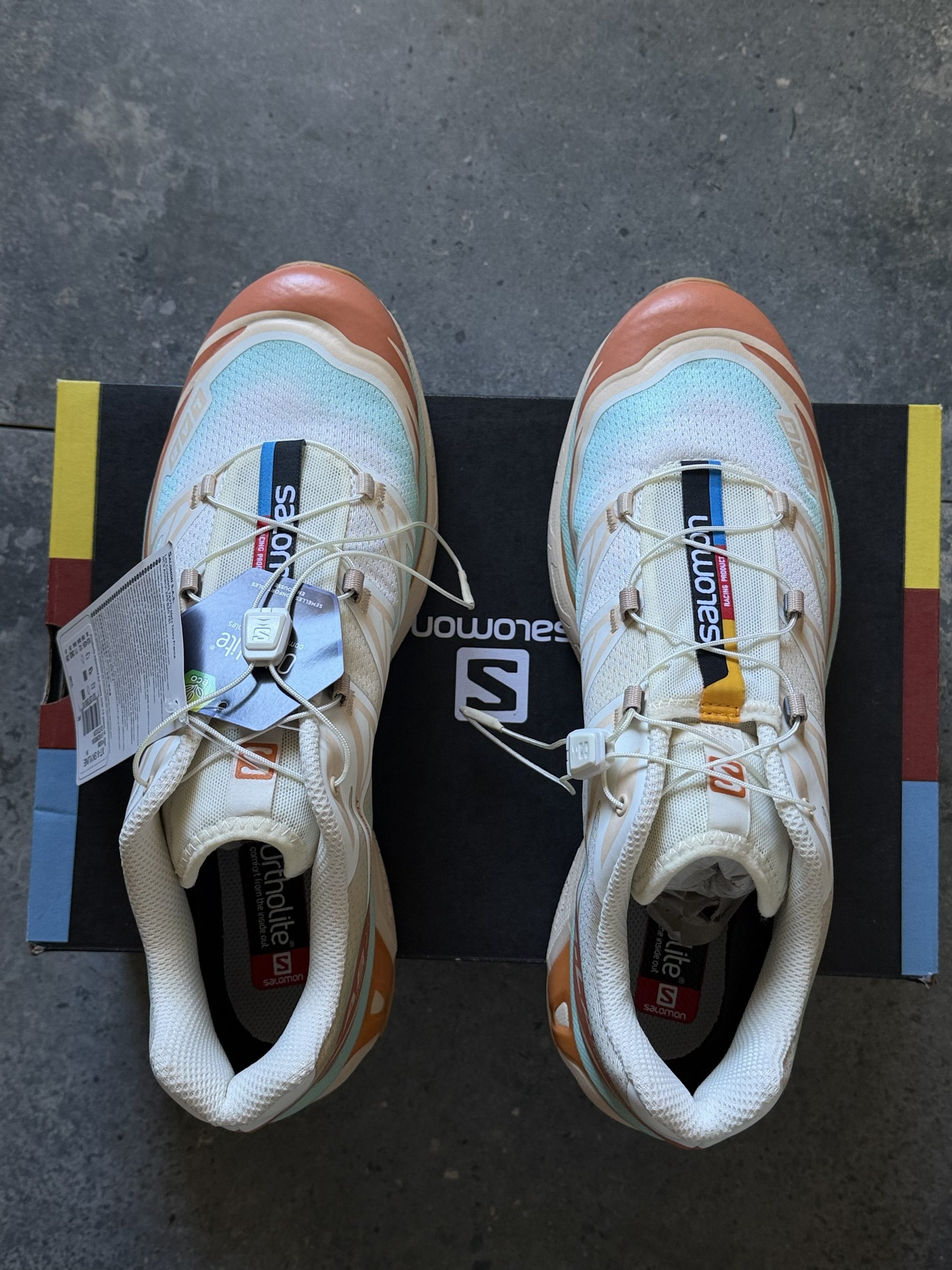 Top-down view of a pair of Salomon running shoes on a black box, beige and peach tones with light blue mesh.