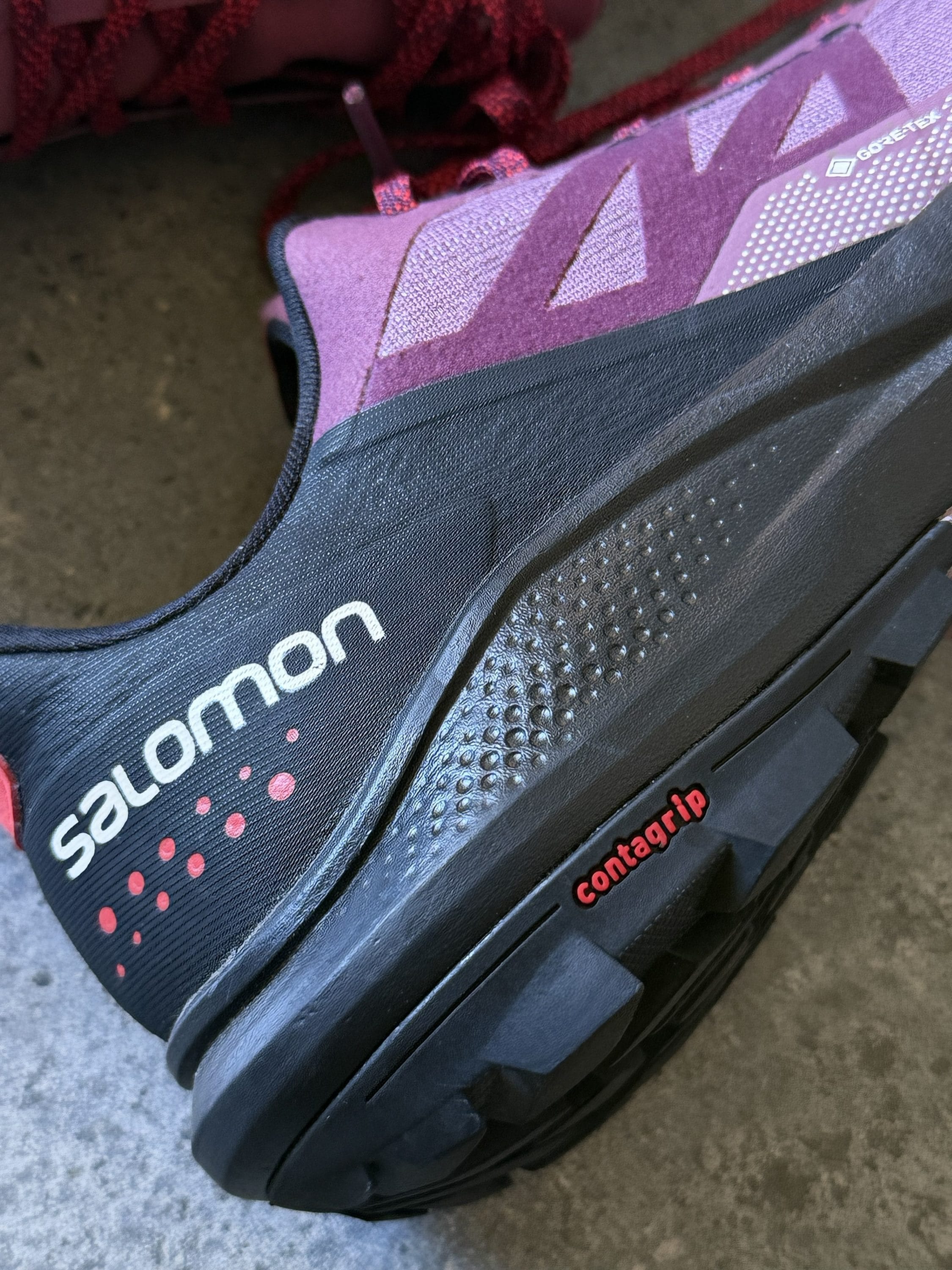 Close-up of a purple-pink Salomon trail running shoe showing the textured upper and Contagrip outsole with red laces visible.