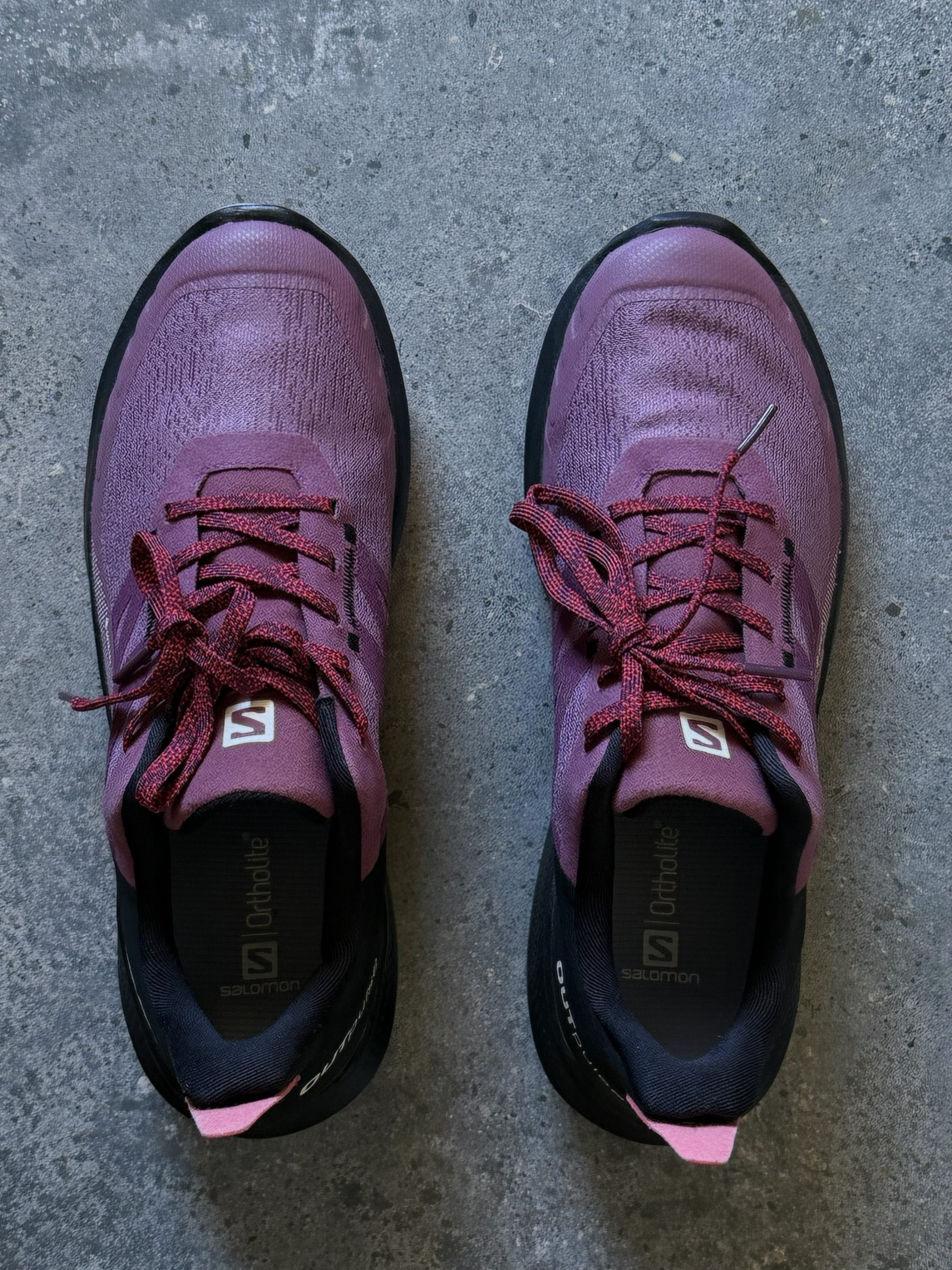 Pair of purple Salomon hiking shoes seen from above on a concrete floor, red laces twisting across the shoes.','Top-down view of two purple sneakers with red laces on a rough gray concrete surface.','A pair of purple athletic shoes placed side by side on a concrete floor.','Close view of purple trail shoes with pink accents and red laces.