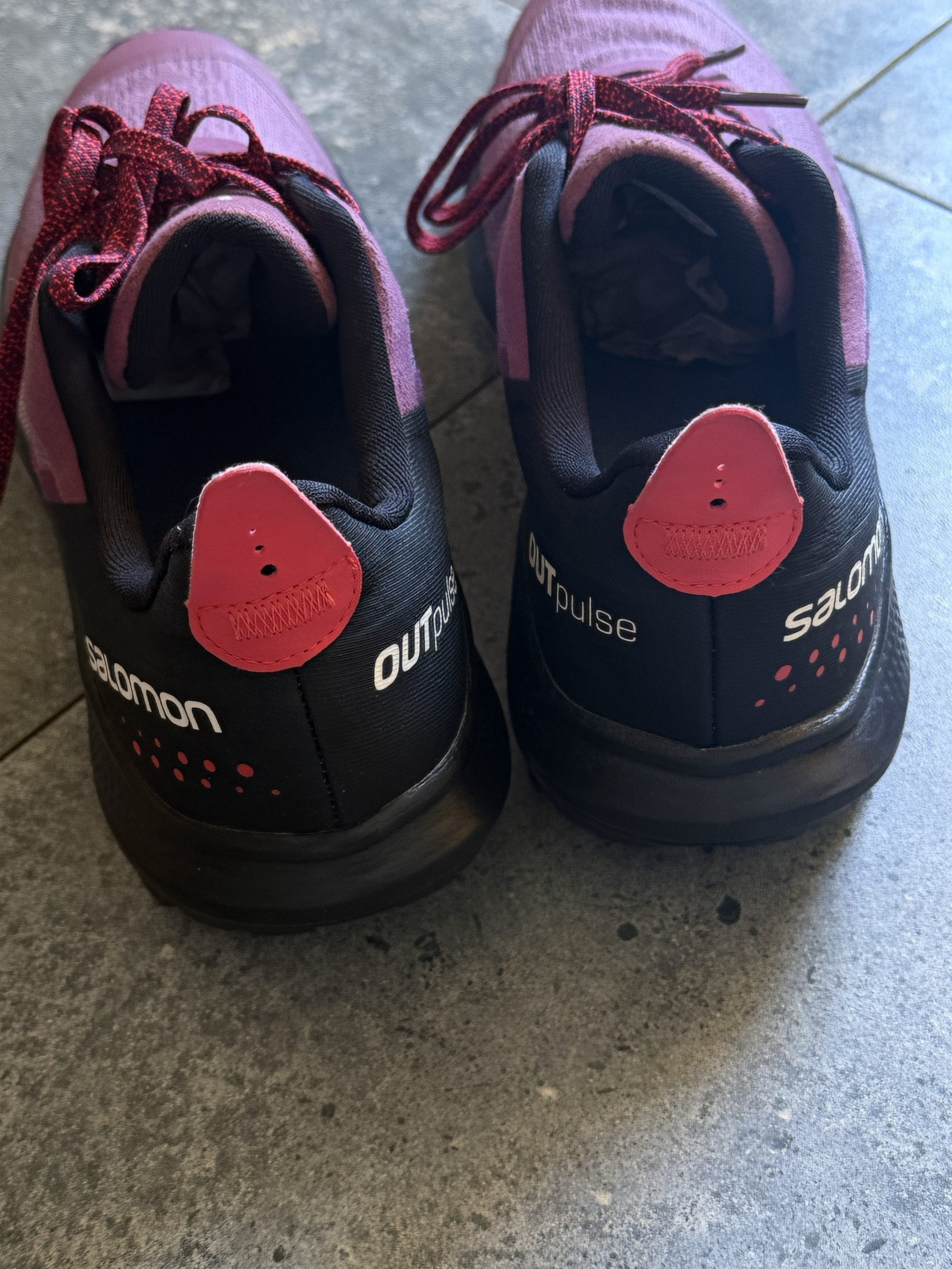 Pair of purple athletic shoes with pink accents viewed from behind on a gray floor, laces loose on top of the shoes.