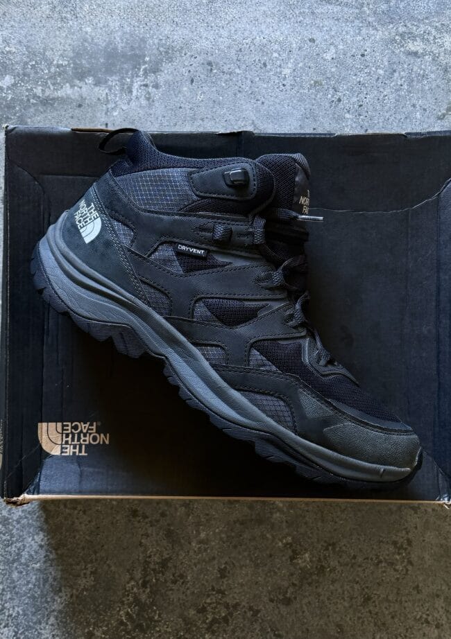 Black The North Face hiking boot rests on its box on a dark concrete floor.