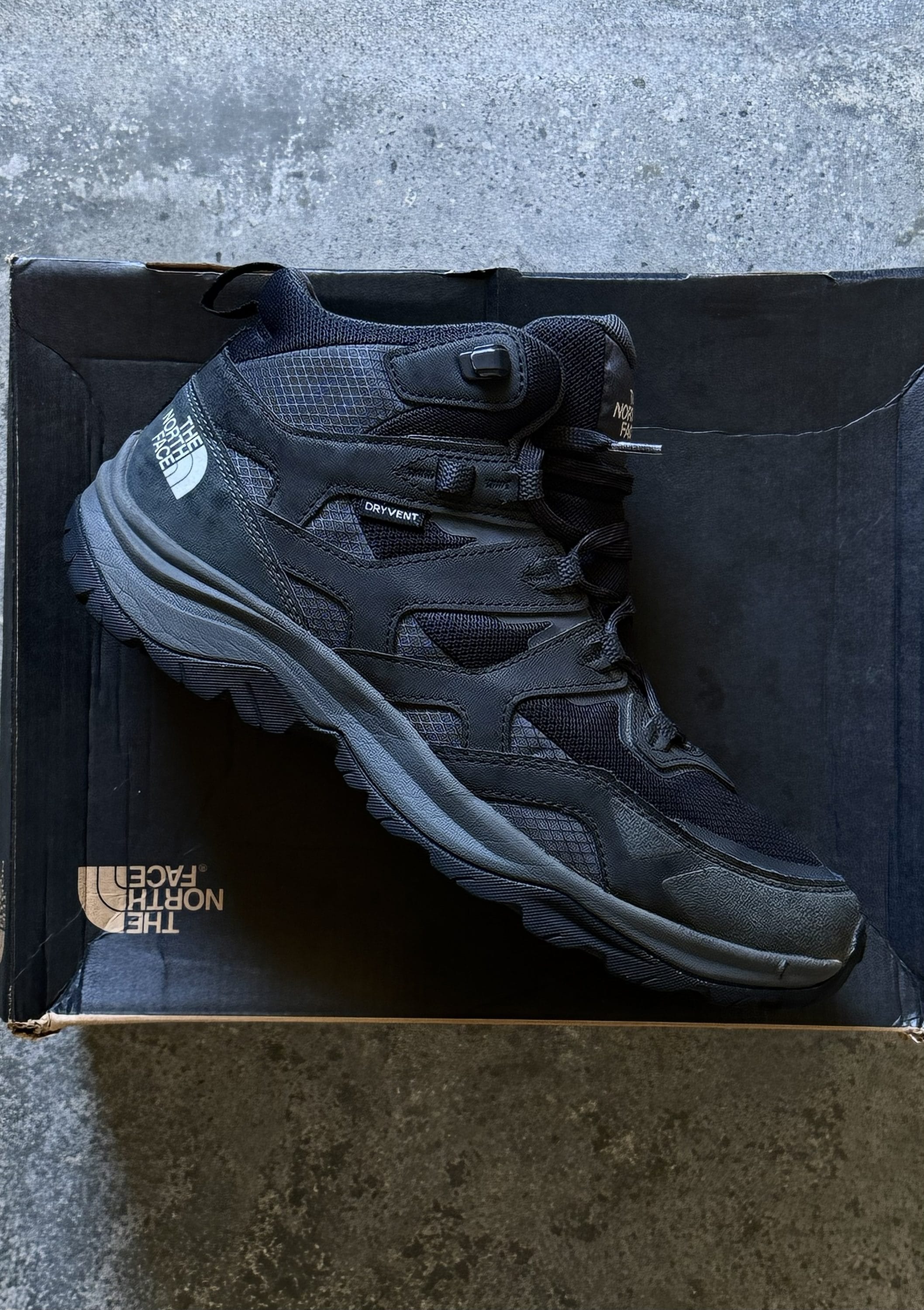 Black The North Face hiking boot rests on its box on a dark concrete floor.