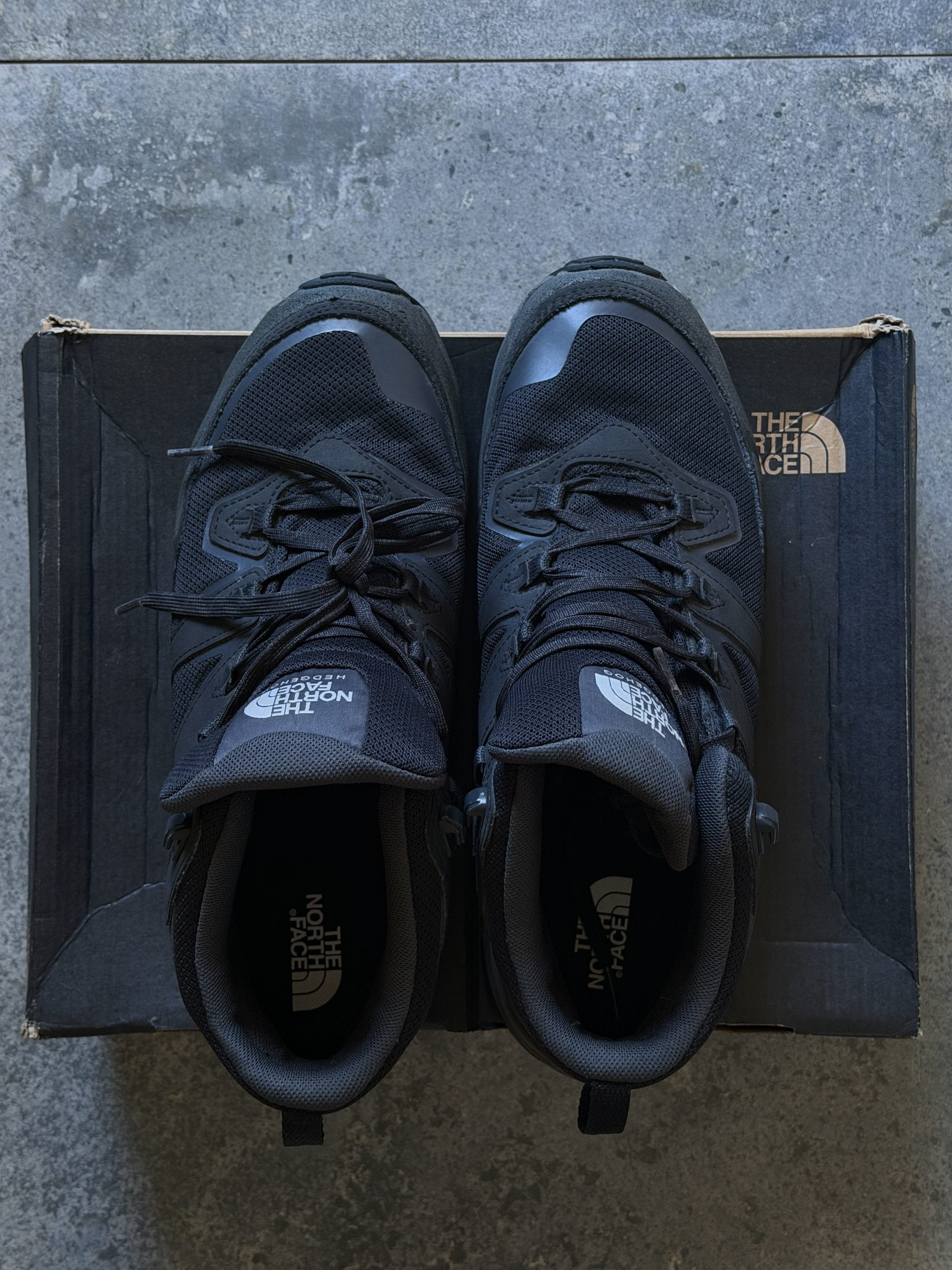 Pair of black athletic shoes resting on a dark shoebox, with The North Face logo visible on the box and tongue labels, on a gray concrete floor.