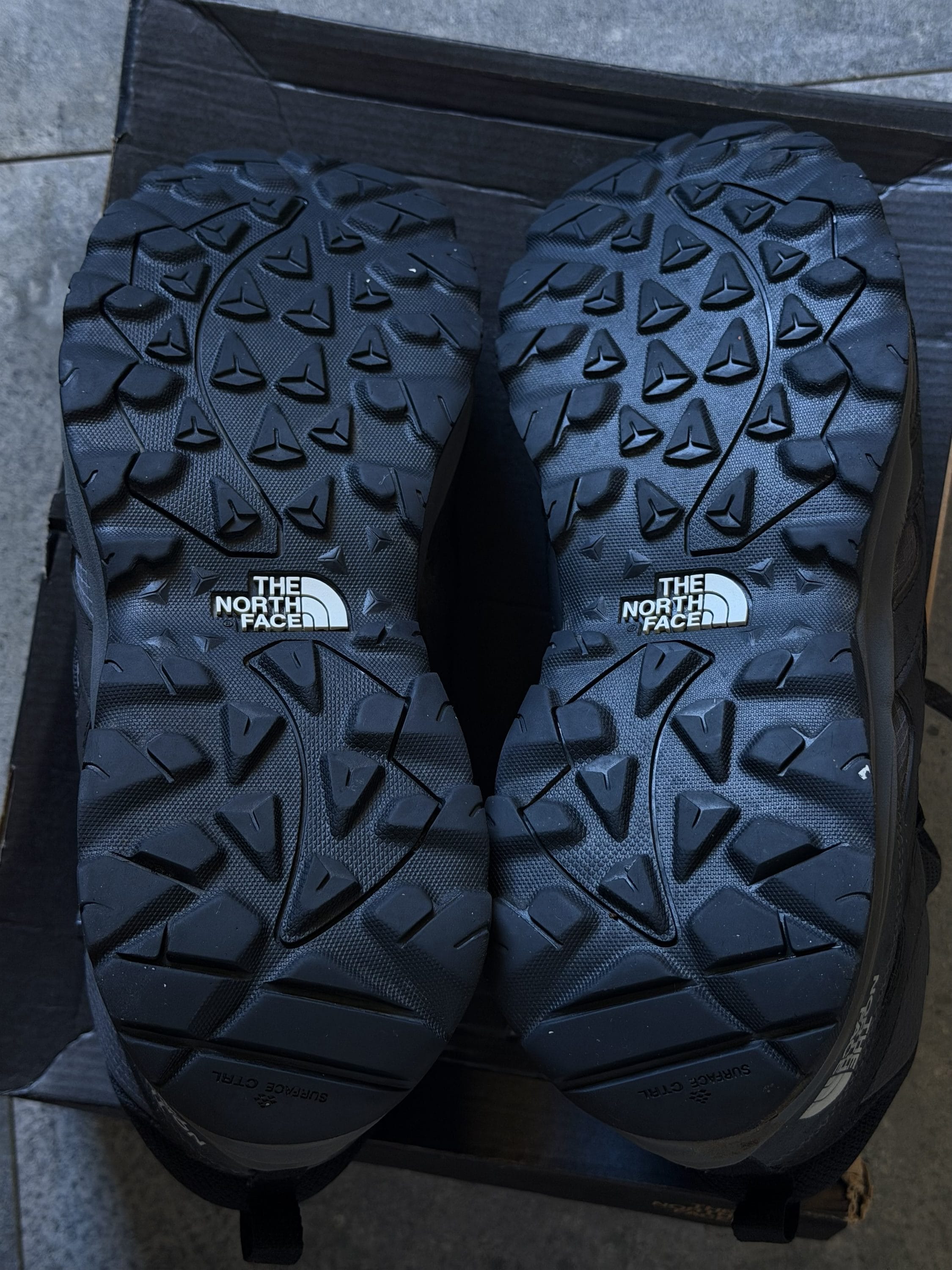 Two black The North Face hiking shoes with pronounced lug tread resting in a box.NOTE: ensure concise phrasing, length around 80 chars for accessibility.