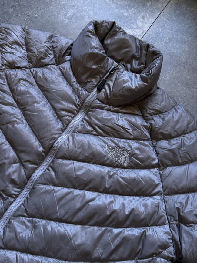 Close-up of a gray The North Face puffer jacket showing quilted sections and a front zipper with a high collar folding over the zipper pull.