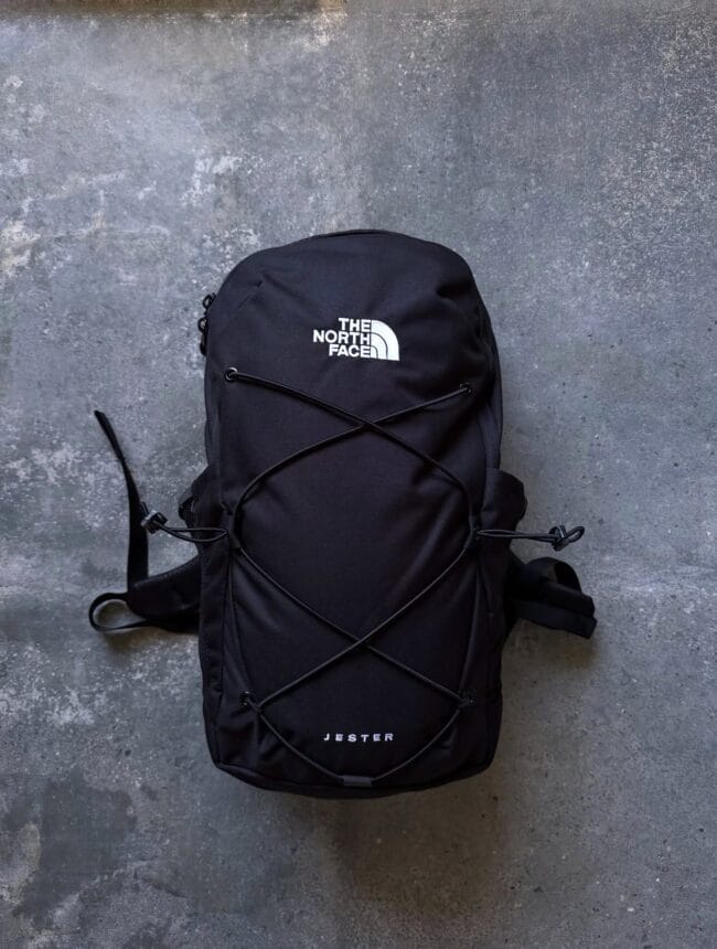 Black The North Face Jester backpack with crisscross bungee cords on a gray concrete floor.