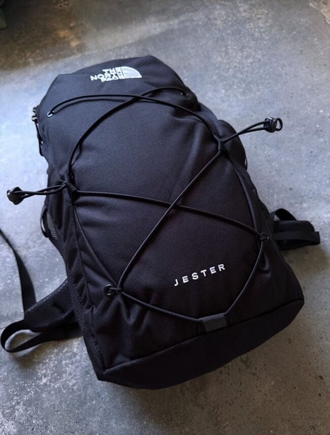 Black The North Face Jester backpack lying on a concrete floor, with bungee cords crisscrossing the front.